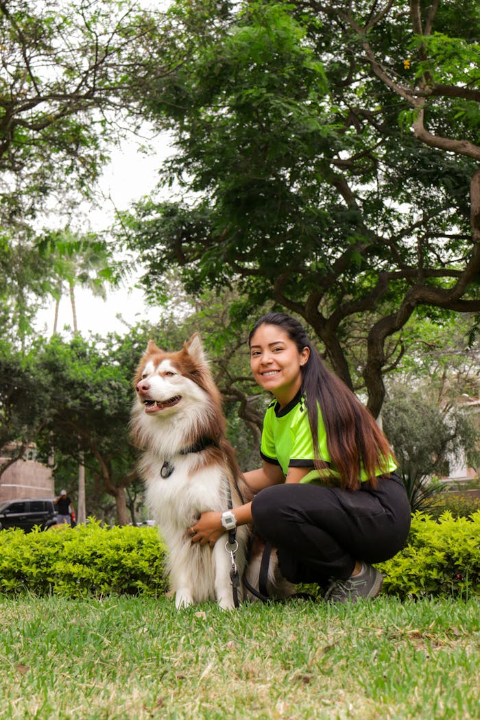 A woman crouches next to her happy Husky in a vibrant green park under a clear sky.